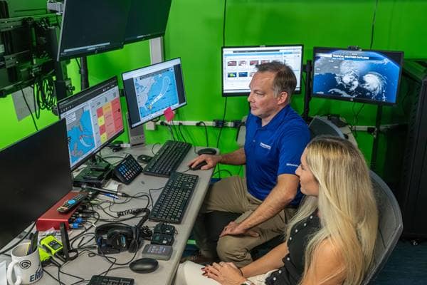 Dr. Rob Eicher and alumna Madison Croker review hurricane forecast graphics in Embry-Riddle’s Weather Broadcast Studio. The graphic to the left shows a new presentation of the cone of uncertainty with a brief chart listing various landfall locations and intensification potential at each.