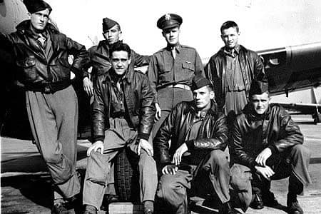Black and white photo shows six men in leather bomber jackets and caps posing in front of a military aircraft with a man in a lieutenant hat, dress shirt and tie.