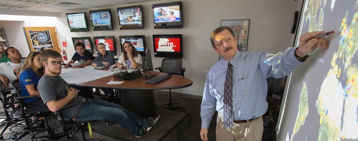 A professor points toward a map of the world while students in front of a wall of monitors look on.