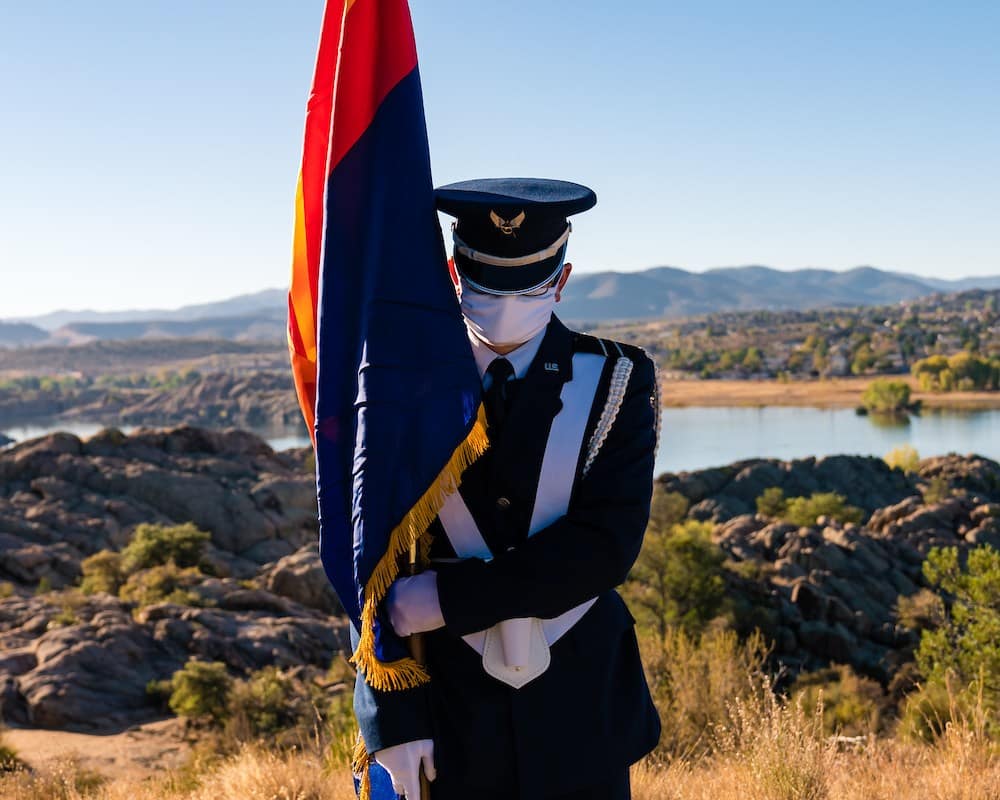 A masked ROTC cadet stands with the Arizona flag in front of Willow Lake