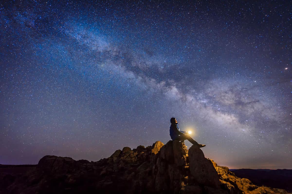 Man sitting on a cliff looking at stars.
