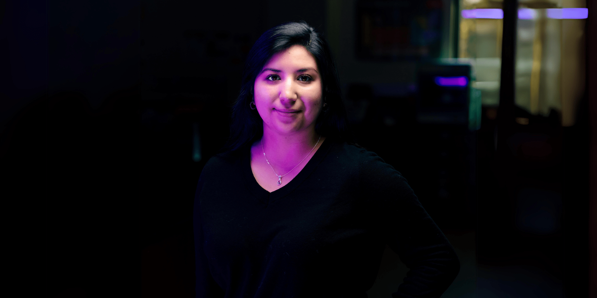 Narayt poses in a dark room with equipment glowing and softly focused in the background.