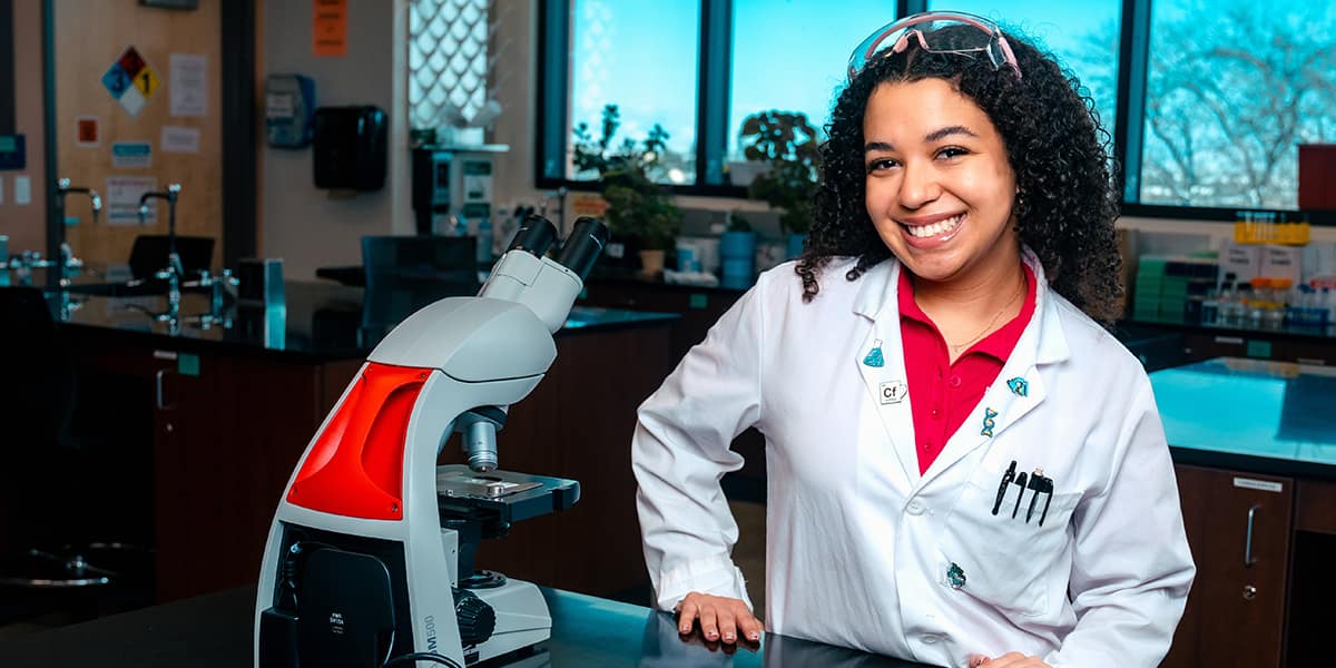 Makenzi, a dark skinned woman with curly hair, smiles in a white lab coat next to a microscope.